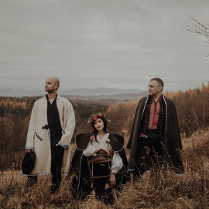 Photo of the band: a woman sitting and two men standing in folk clothes; mountainous landscape in the background