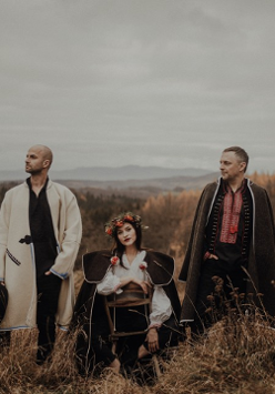 Photo of the band: a woman sitting and two men standing in folk clothes; mountainous landscape in the background