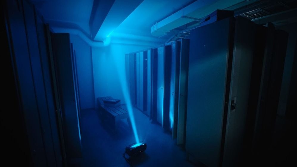 The room resembles a locker room or a changing room at a gym, it is dark, there is a device on the floor from which a narrow, blue beam of light shines towards the ceiling.