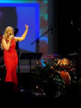 Photo of the woman singing to the microphone on a stage. On the right - percussion instruments.