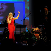 Photo of the woman singing to the microphone on a stage. On the right - percussion instruments.