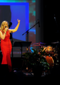 Photo of the woman singing to the microphone on a stage. On the right - percussion instruments.