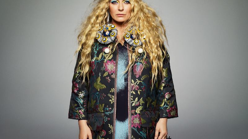A picture of the artist - a woman with long, blond, wavy hair, dressed in a colourful long jacket and a black hat who is looking into a camera. Grey background.
