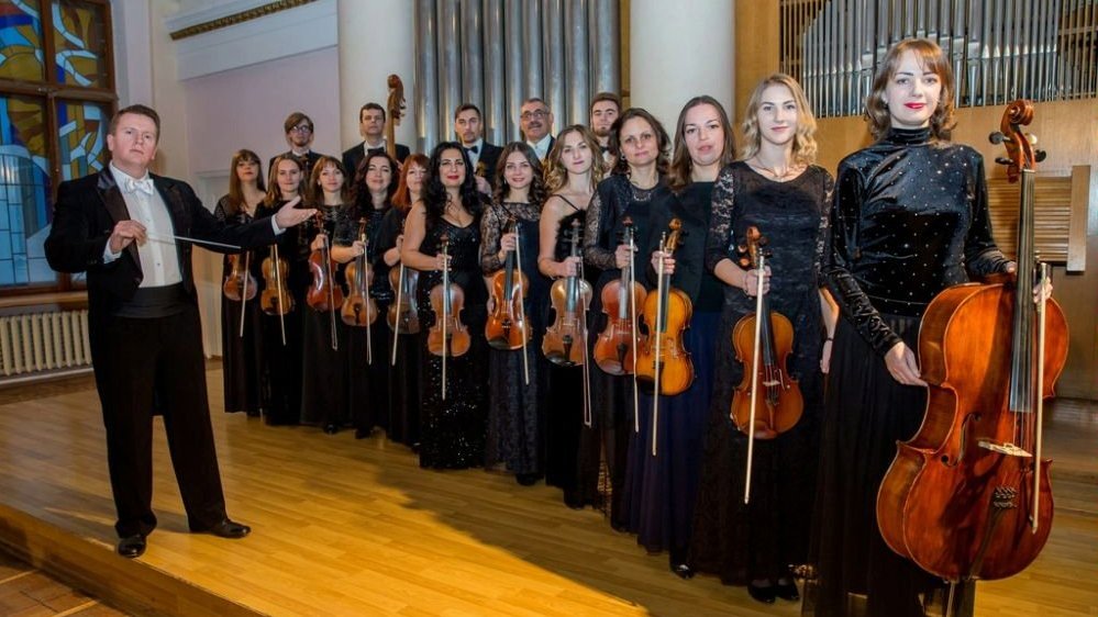 Photo of the orchestra - a group of musicians standing in two rows holding their string instruments; on the left - the conductor.