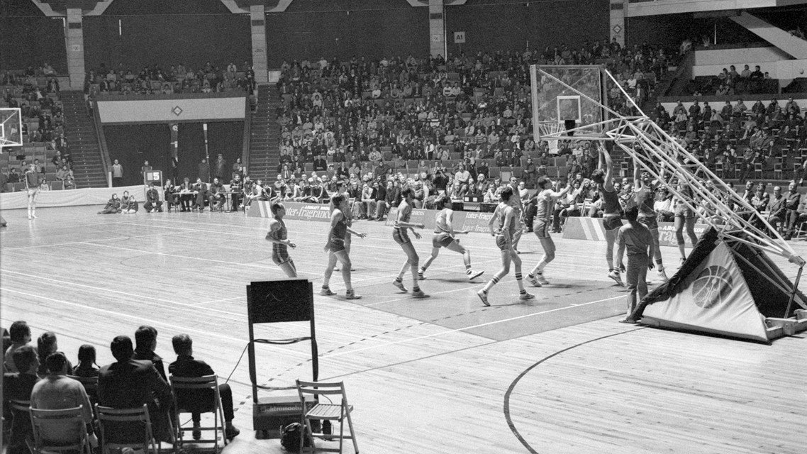 Black and white photo of a basketball game in a sports hall. Tribunes with fans around the pitch.