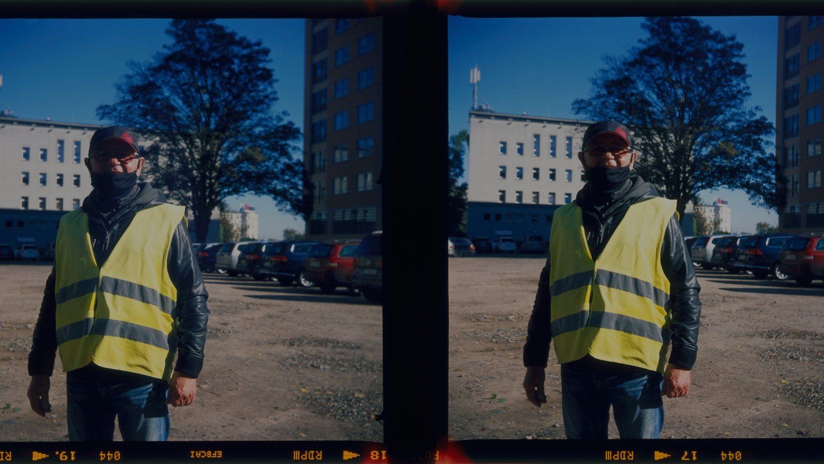 Stereoscopic photograph presenting a man dressed in a reflective vest; the man is standing on a car park. As a background - a few parked cars in a row and two buildings.
