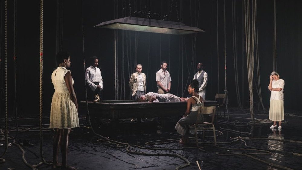 A scene from the performance: a person lying on a big table and some people standing around a table, one woman sitting on a chair.