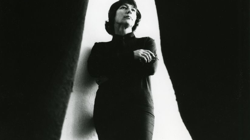 Black and white photo of Magdalena Abakanowicz - a lady dressed in black dress with her arms folded on white background. On the left and right - black curtains