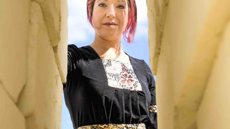Photo of a woman dressed in colorful clothes, who is looking straight into the camera, peering into a narrow crack between two walls. Blue sky as a background.
