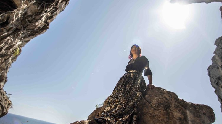 Photo of a woman in a long patterned skirt, who is sitting on a rock. A fragment of a rock in a foreground and the blue sky and sea as a background.