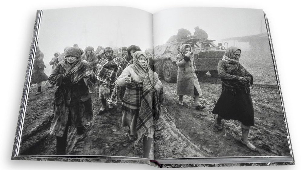 The inside of a book with black and white photo. In the photo marching women, some of them covered with blankets; in the background a battle tank.