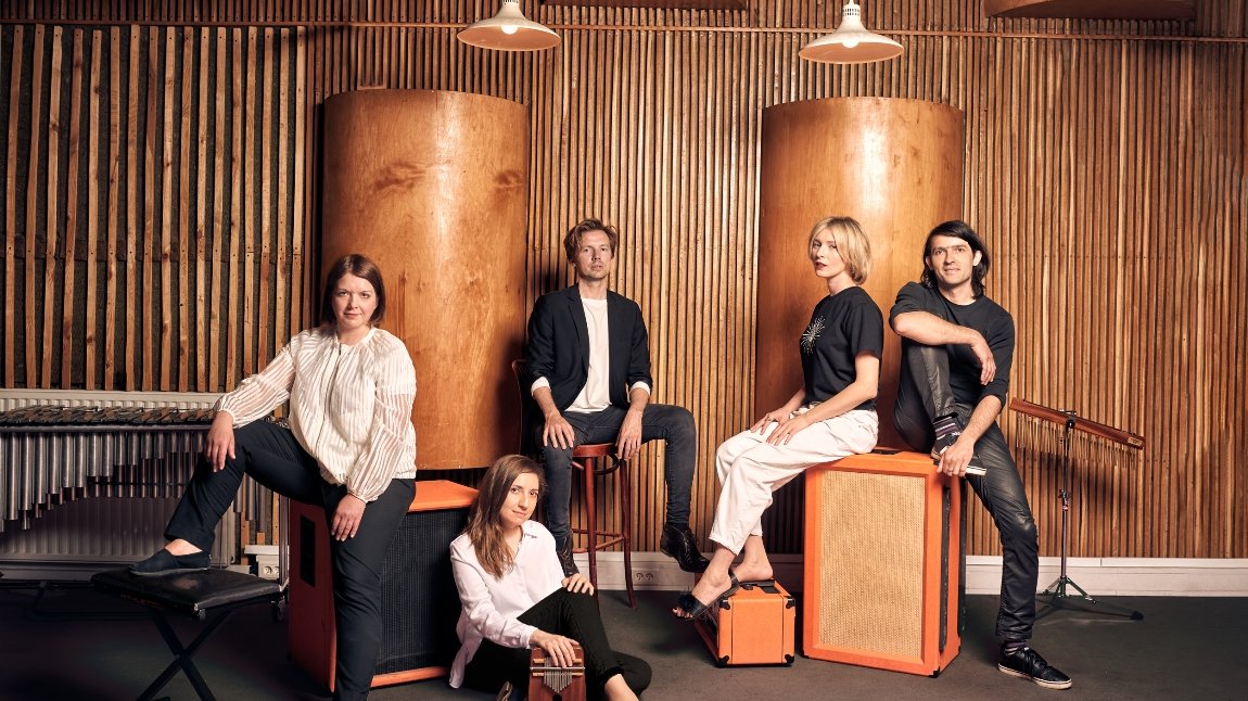 Picture of a band: three women and two men - four of them sitting ond chairs and a box that look like a loudspeaker, one woman is sitting on a floor. The brown-orange wall with two columns in a background.