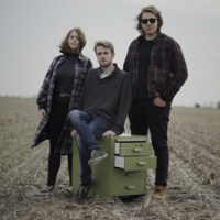 Photo of the band - two men and a woman on an empty field by a small chest of drawers.