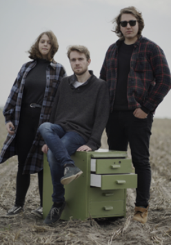 Photo of the band - two men and a woman on an empty field by a small chest of drawers.