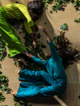 Two dancers - one in green, another in blue clothes - lying on the floor covered with green leaves.
