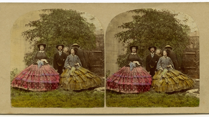 Stereoscopic photograph presenting two women in crinolines and a man in a suit between them. Behind them a man and a woman turned backwards. A tree in a background.