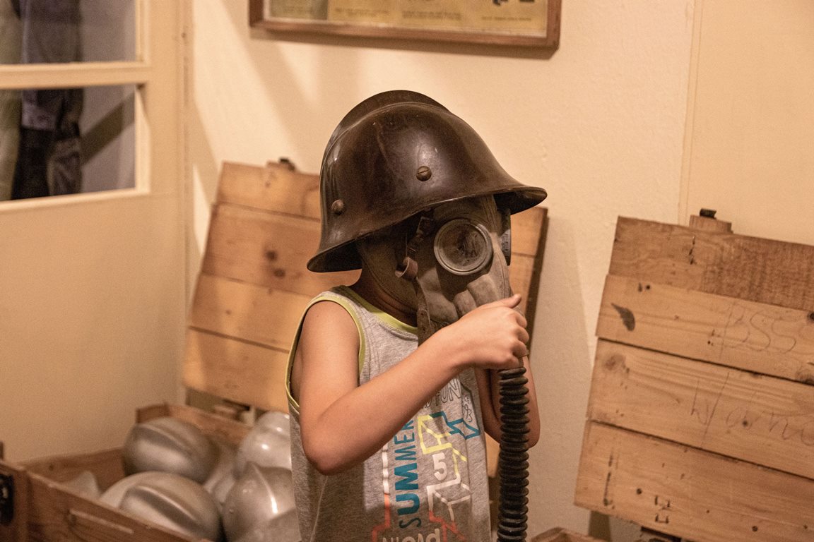 The picture presents a child in a gas-mask and a helmet. Wooden open chests in the background. - grafika artykułu