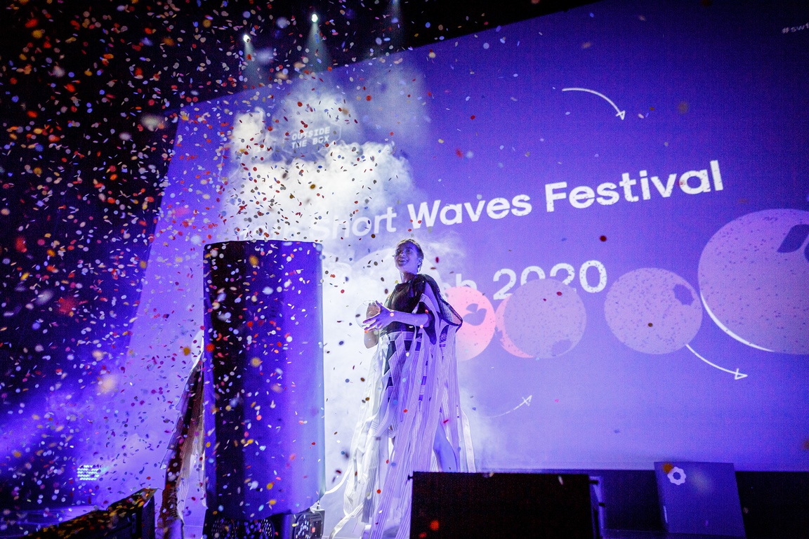 A woman standing on a stage. A big screen in the background. The picture in blue and violet colours, in front of the stage spread colourful confetti. - grafika artykułu