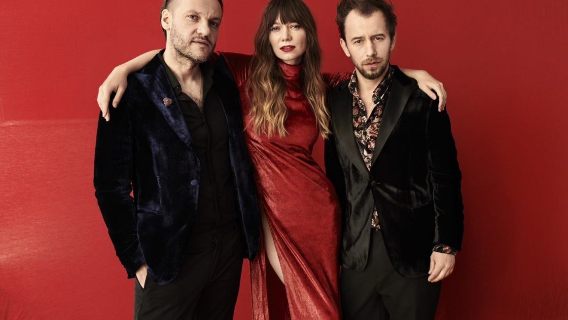 A picture of one woman and two men on the dark red background. A woman in the middle puts her arms around two men