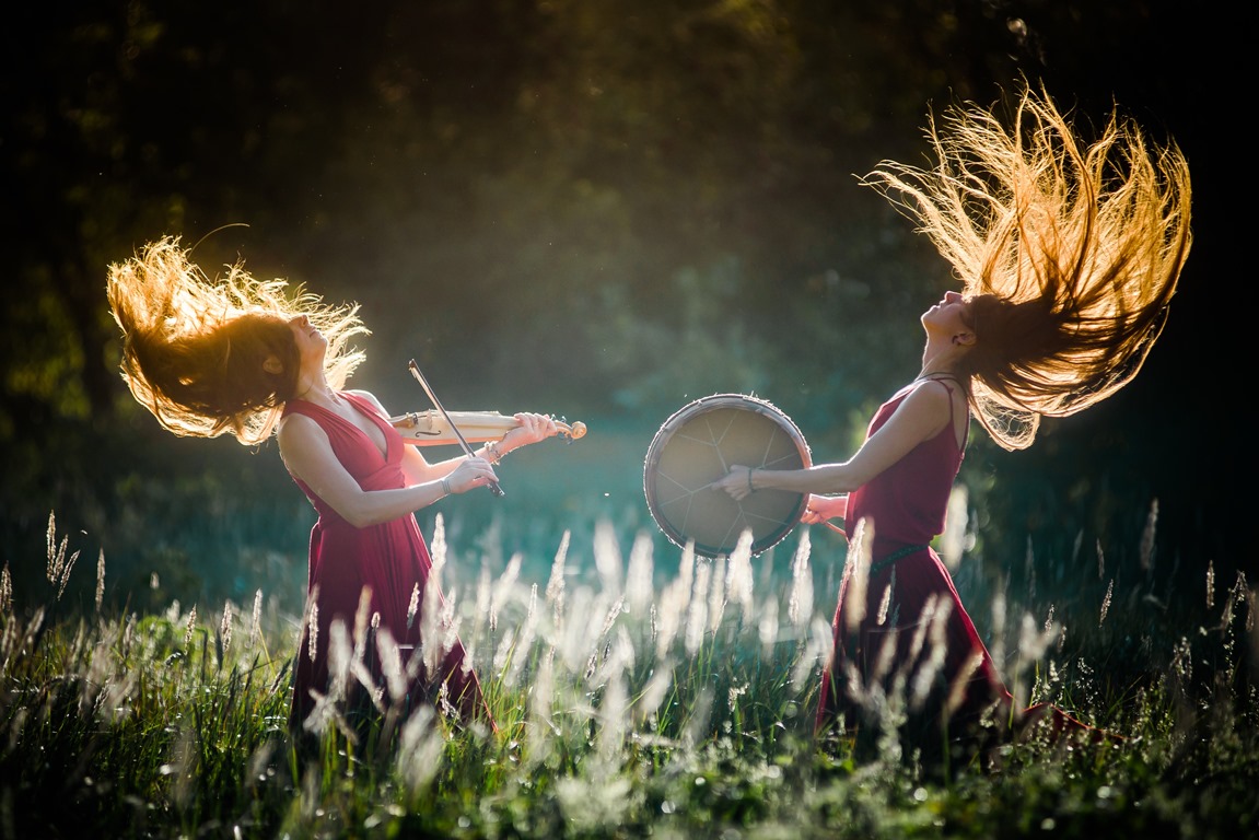 The picture presents two women in red dresses, with music instruments in their hands. They are standing in the meadow opposite each other in artistic trance - grafika artykułu