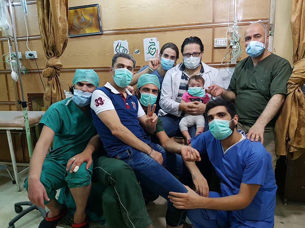 The picture presents a group of 7 men in a medical care room. They are in medical garments with masks on their faces. One man is holding a little child on his knees. - grafika artykułu