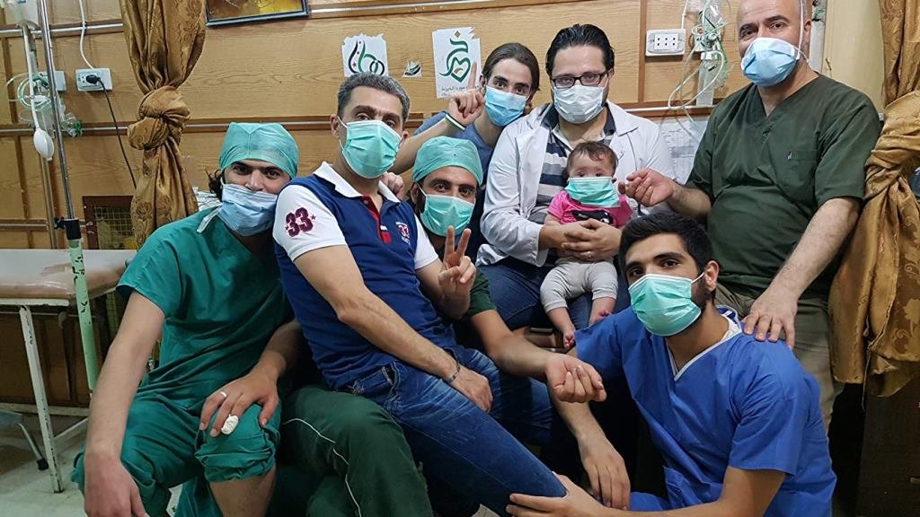 The picture presents a group of 7 men in a medical care room. They are in medical garments with masks on their faces. One man is holding a little child on his knees.
