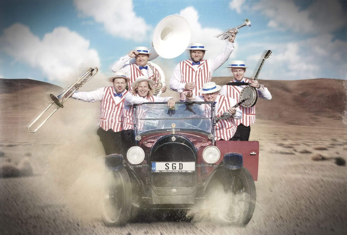 Photo of 6 men in an old car on a desert, dressed in white shirts, white and red vests and white hats, holding musical instruments in their hands. - grafika artykułu
