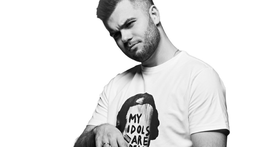 Black and white photo of Łukasz Ojdana, who is sitting on a chair and gesturing with his hands. The man is looking at the camera.