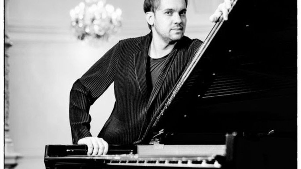 Black and white photo of the artist standing behind the grand piano