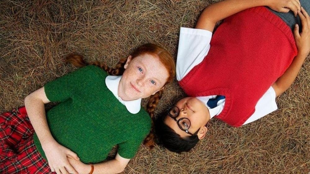 A girl with red hair and a boy in glasses are laying on the ground covered with hay. The boy is looking at the girl, the girl is looking at the camera.