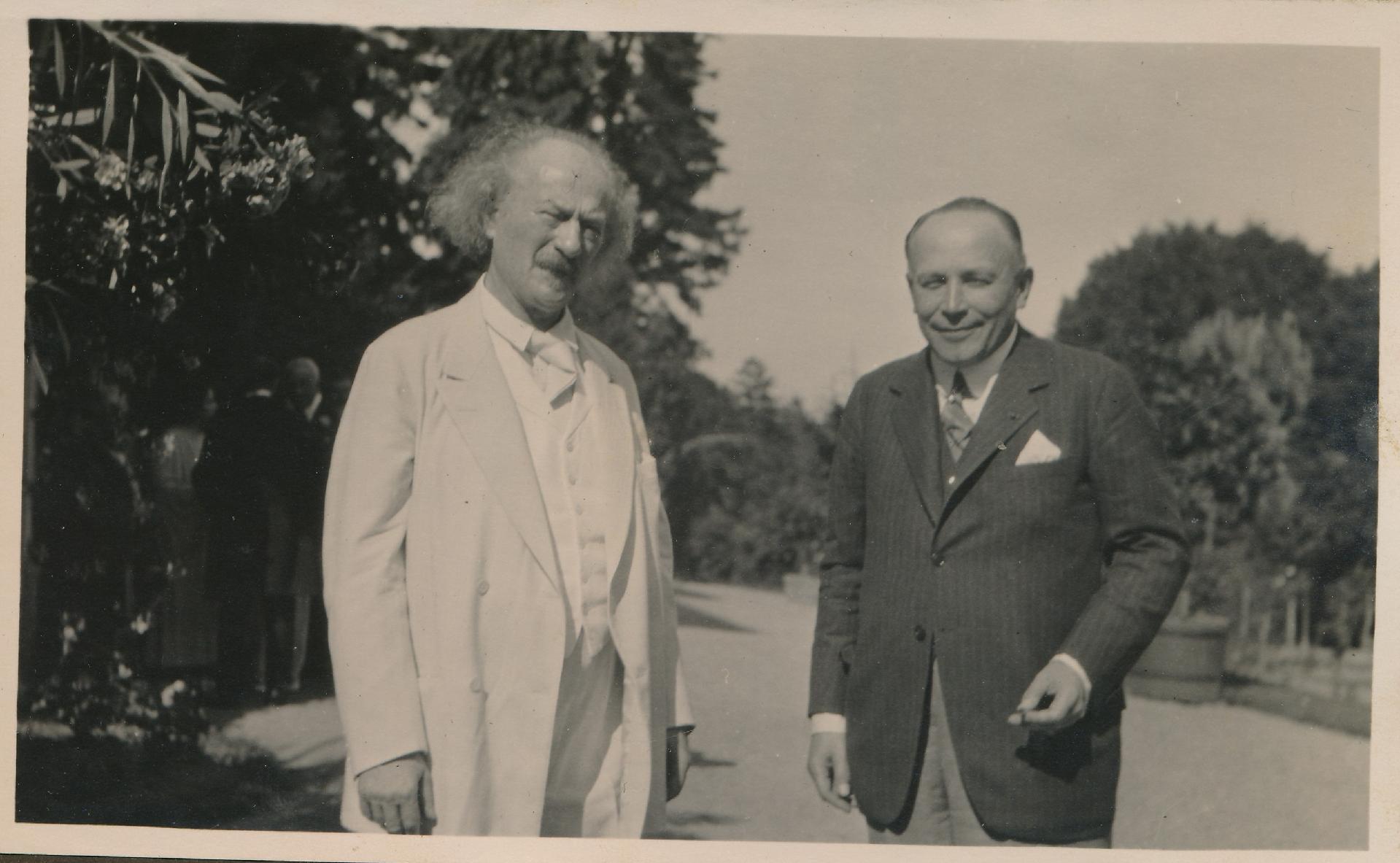 Black and white picture of two elegantly dressed men (one of them Ignacy Jan Paderewski) standing on the road, trees in the background. - grafika artykułu