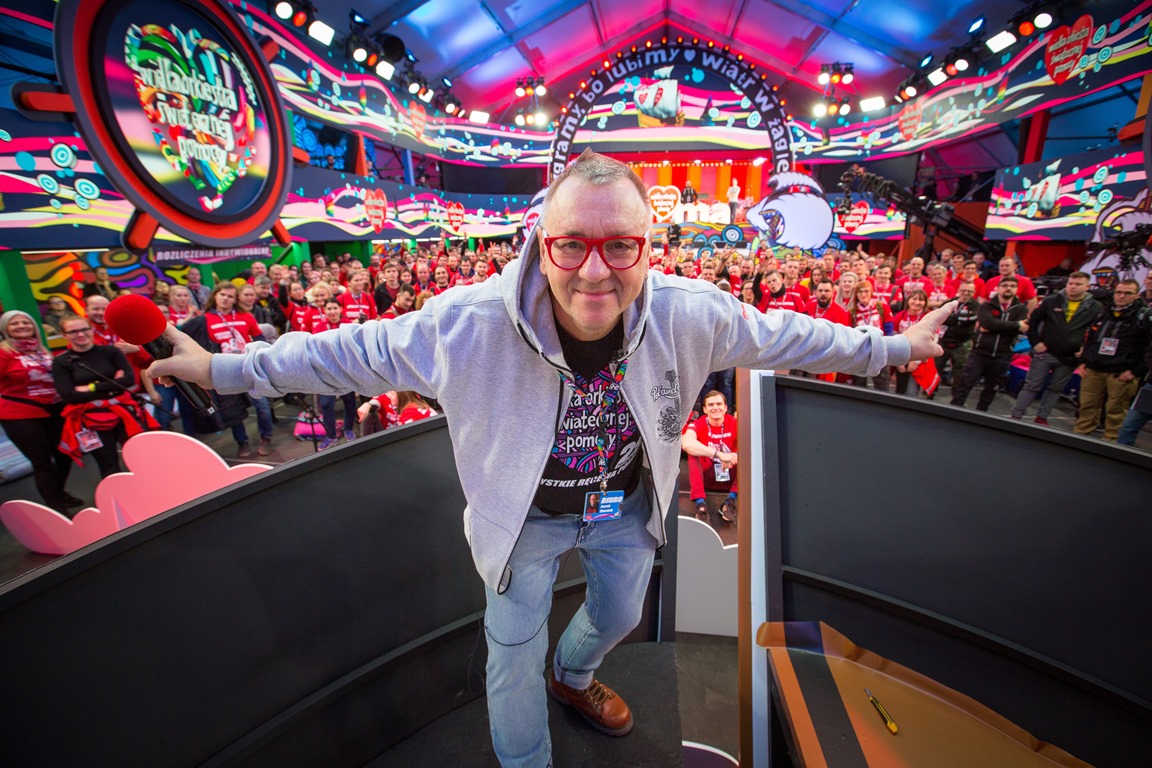 Colourful photo of Jurek Owsiak - creator of Grand Orchestra of Christmas Charity, taken in a TV studio. In the background people mostly dressed in red t-shirts with Grand Orchestra of Christmas Charity logo. - grafika artykułu