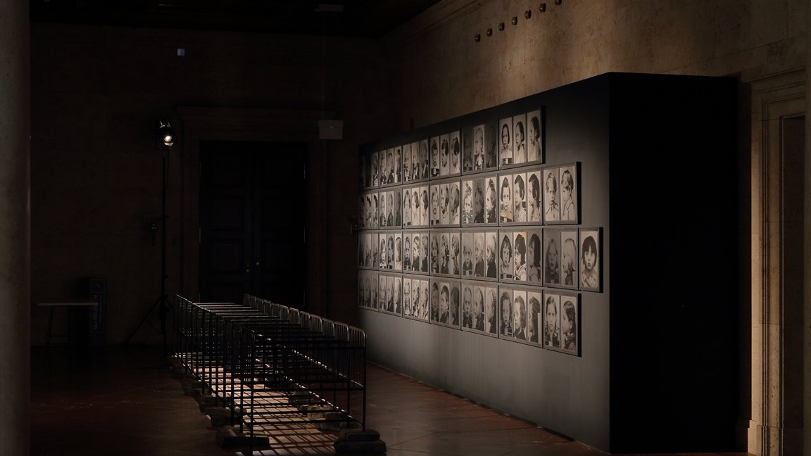 A photo from the exhibition: a dark room with black and white portraits of children on a wall. In the middle of the photo old fashioned metal baby beds.