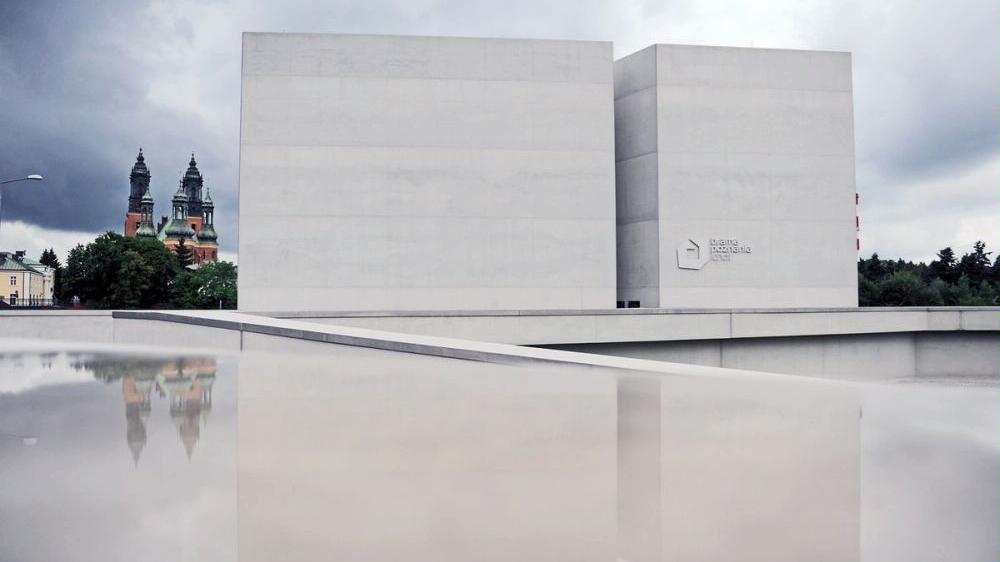 In the foreground concrete cube-shaped building without windows, in the background the building of Poznań's Cathedral and some trees. - grafika artykułu