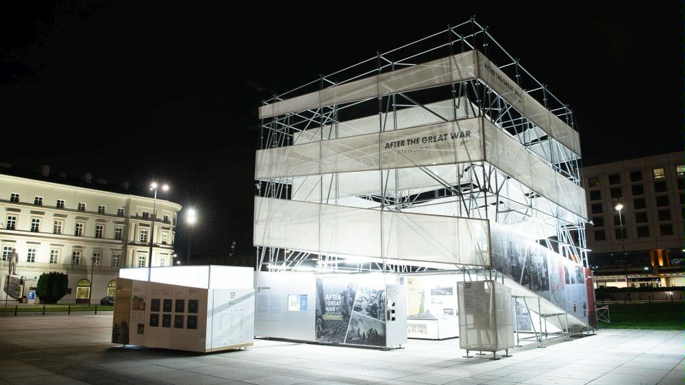 The photo shows a metal cube-shaped illuminated structure with inscriptions and photos. Photo taken at night, an illuminated building in the background - grafika artykułu