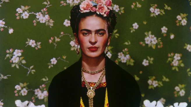 Portrait of Frida Kahlo - a dark-haired woman dressed in dark clothes, wearing golden necklace and earrings, with flowers in her hair. Dark green wallpaper with fair flowers as a background.