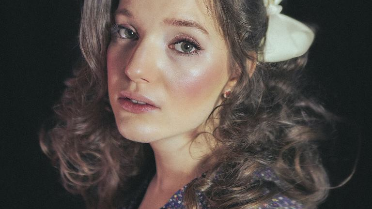 A picture of the artists face - a young woman in dark curly hair with a bow in it.