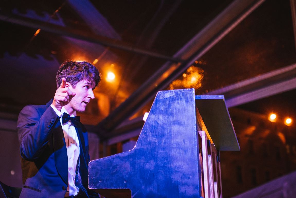 Photo of a man in a suit sitting at the piano and pointing at something with the finger of his right hand - grafika artykułu