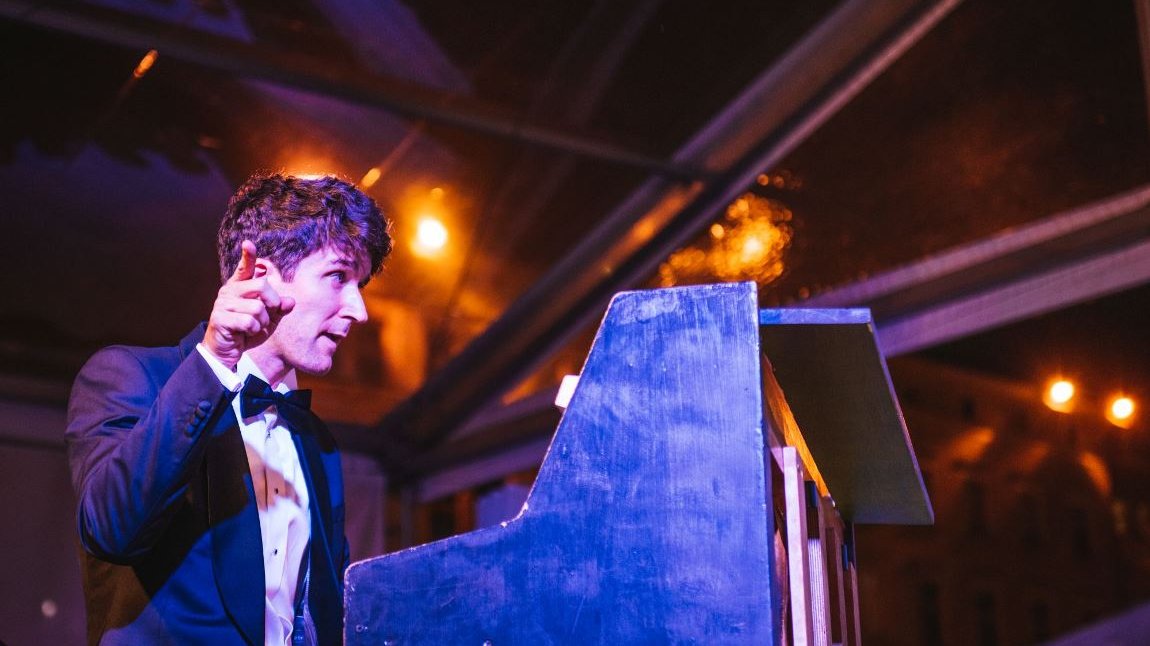 Photo of a man in a suit sitting at the piano and pointing at something with the finger of his right hand