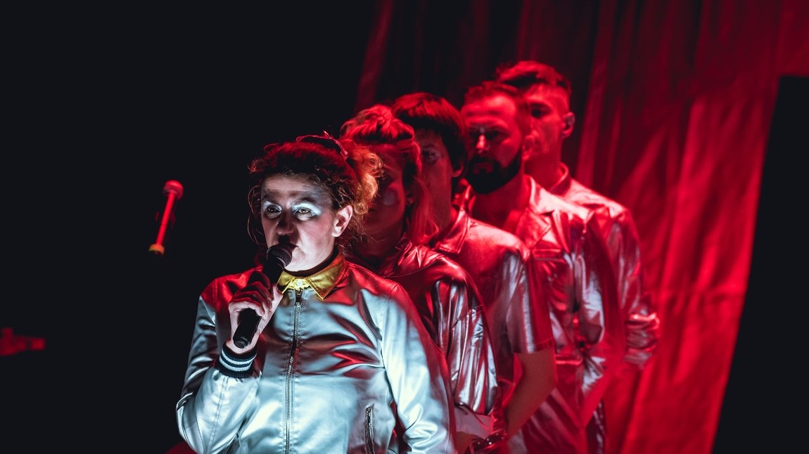 Picture from the performance: 5 people standing in a row. The first one is wearing silver clothes and is holding a microphone to which she is speaking. The other people lighted by red light.