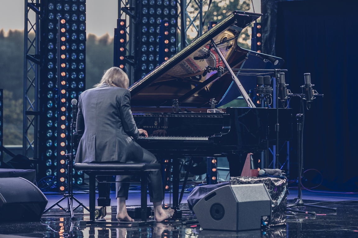 Photo of Leszek Możdżer sitting back to the camera and playing piano on an open-air stage - grafika artykułu