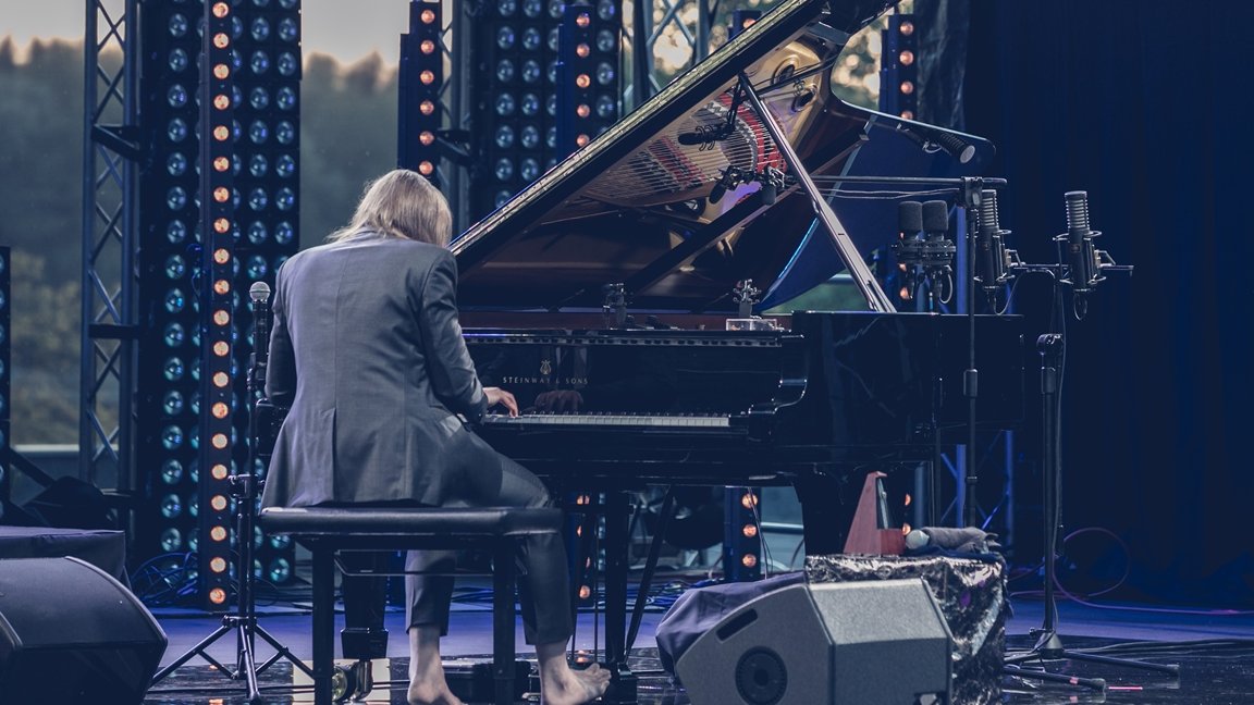 Photo of Leszek Możdżer sitting back to the camera and playing piano on an open-air stage