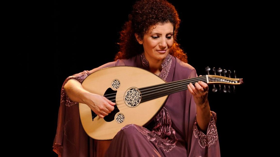 Photo of a woman in a long dress. The artist sits and holds an oval stringed instrument in her hands