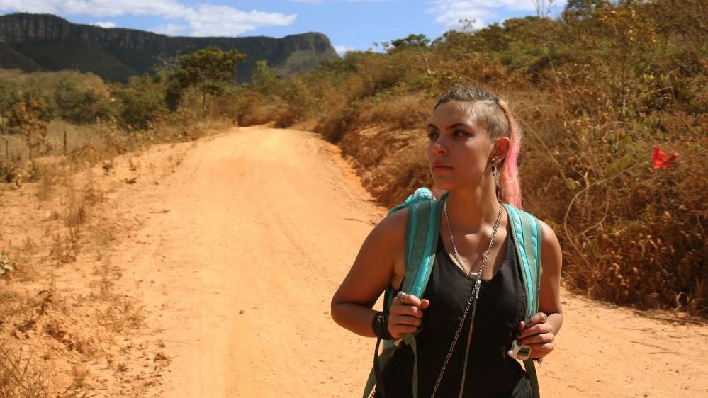 Photo from the movie: a woman with a rucksack on her back marching along a sandy way. Some bushes and grass in the background
