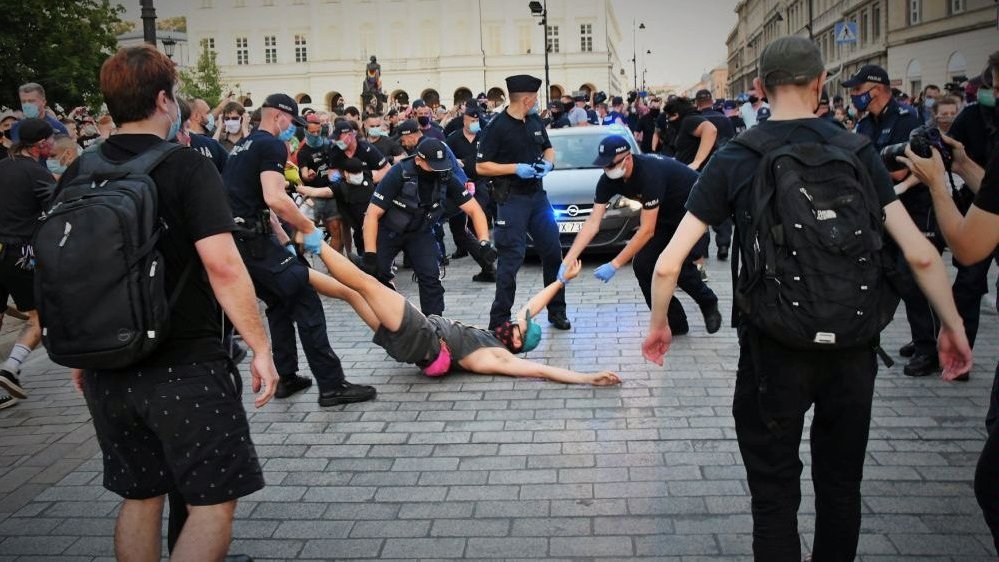 Photo from the movie: policemen pulling a young man, who is lying on a pavement. In the background a car, policemen and other people.