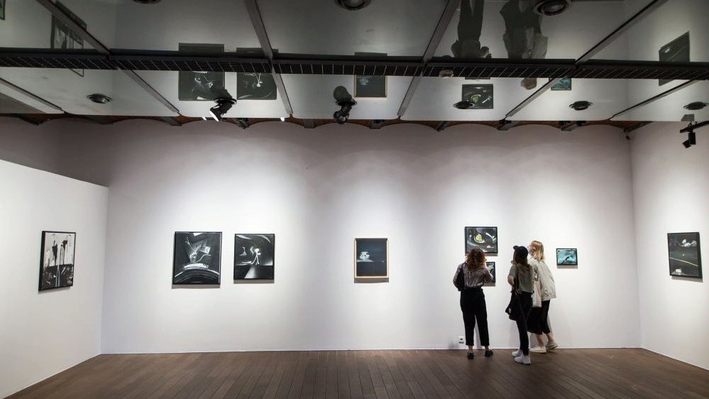 Exhibition room - Noriaki's works on white walls. Three people who are looking at the pictures.