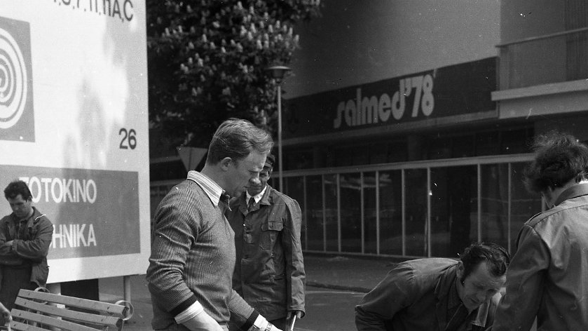Black and white picture of a few men who are renovating a bench - one of them is paining it, one is standing by a bench, three other men are pouring the paint from big to small can.