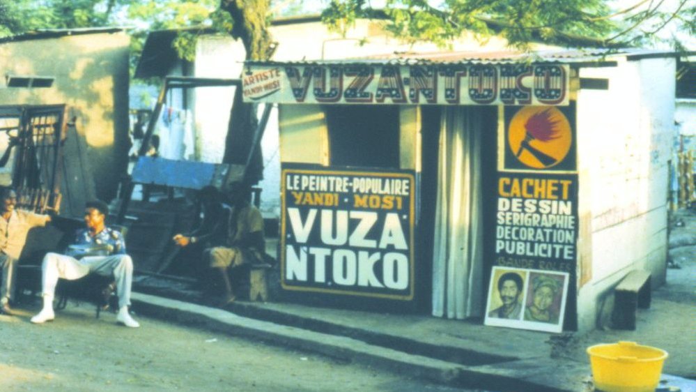 Picture of a man sitting in front of painter's atelier - a small building with signboards