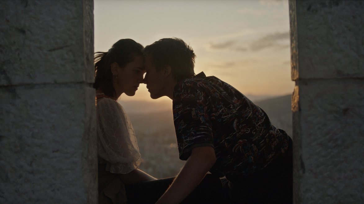 Picture from the movie - a teenage girl and boy touching with their foreheads, between two stone walls at dusk.