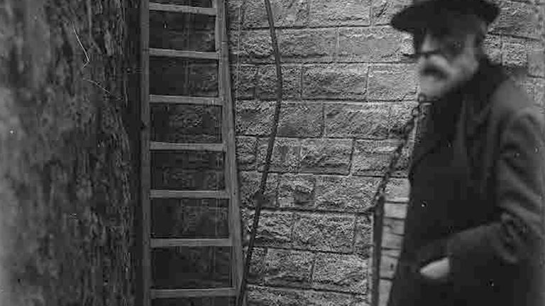 Black and white picture of Antoni Gaudí. Gaudi stands on the right with his hand in a pocket. On the left - a ladder leaning against a wall. A brick wall as a background.